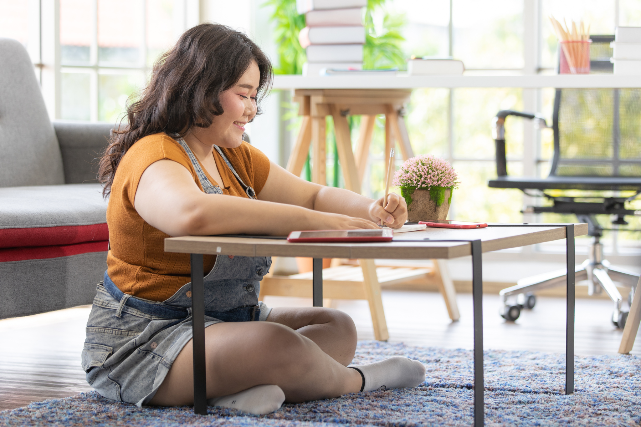 woman with dark hair sits on floor writing, wearing cute overall outfit